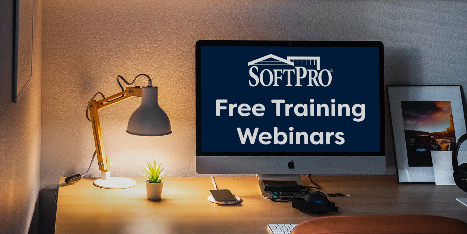 A computer monitor on a desk lit warmly by a lamp beside it; text on the screen reads 'SoftPro Free Training Webinars' in white on a navy blue background